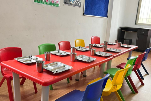 A school dining hall with a long red table, colorful chairs, and stainless steel meal trays prepared for students.