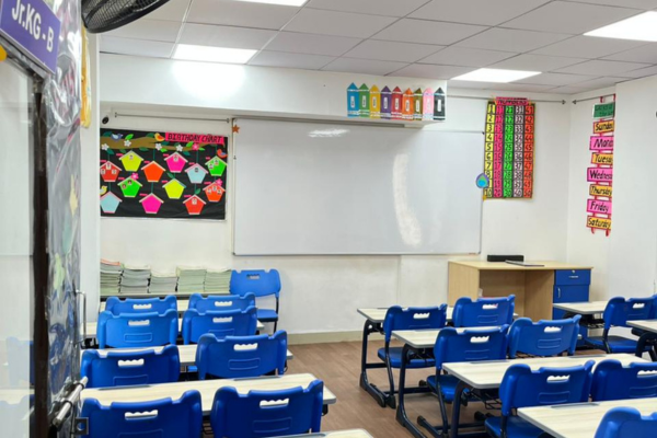 A clean, organized junior kindergarten classroom with blue chairs, white desks, and educational charts on the wall.