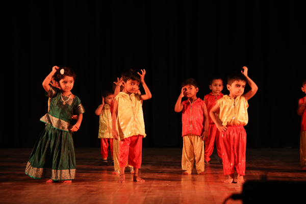 A group of young children from "The Learning Campus" performing a dance routine on a dark stage. The children are dressed in vibrant, traditional Indian-style costumes including green, gold, and red outfits.
