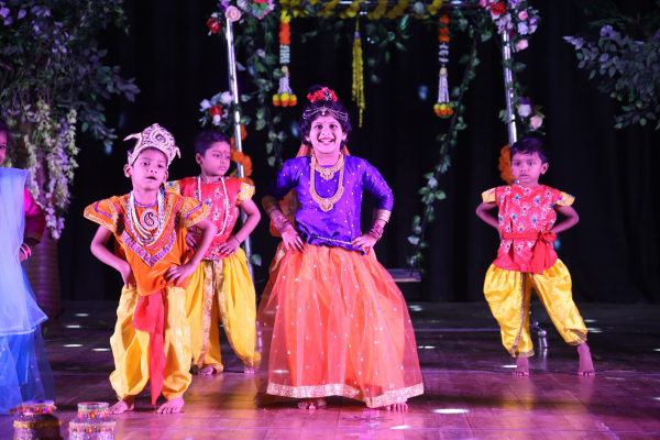 A group of young children from "The Learning Campus" performing a dance routine on a dark stage. The children are dressed in vibrant, traditional Indian-style costumes including green, gold, and red outfits.